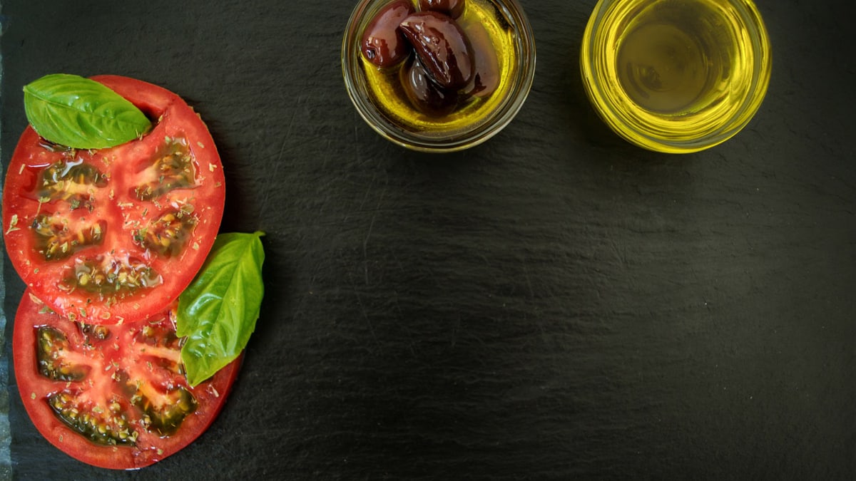 Top view of fresh basil, sliced tomatoes, olives, and olive oil on slate, perfect for Italian cuisine themes.
